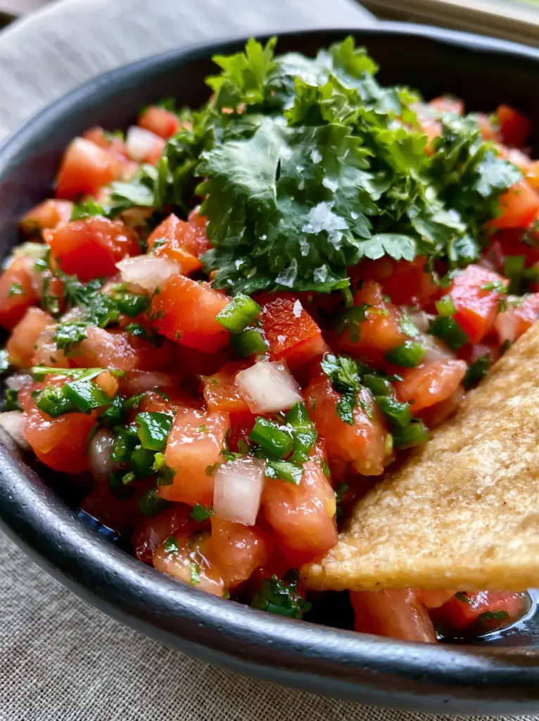 A close-up view of hand-torn cilantro leaves and flaky sea salt over a fresh Pico De Gallo.
